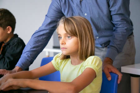 Serious Caucasian girl sitting at table in classroom, reading text on screen or watching video presentation. Unrecognizable teacher standing behind her. Knowledge and digital education conceptの写真素材