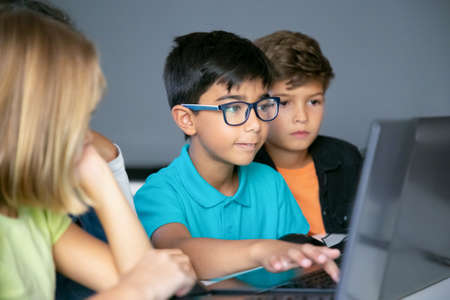 Asian boy typing on laptop keyboard and classmates sitting at table, watching him and doing task together. Pupils using computers during study. Childhood, communication and digital education conceptの写真素材