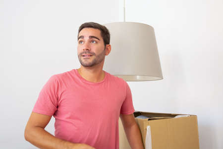 Happy young Hispanic man unpacking things in his new flat, standing near carton boxes, looking away....