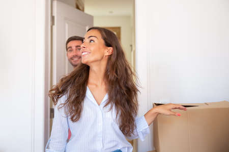 Joyful young man and woman walking through doorway and looking around in their new apartment with carton boxes. Medium shot. New home or surprise conceptの写真素材