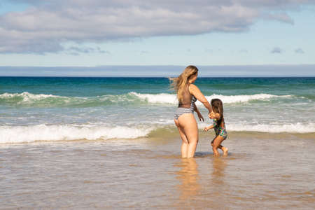 Sweet young mom and little daughter standing ankle deep in sea water, spending leisure time on beach...