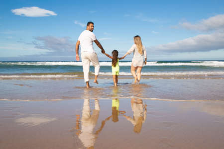 Family couple and kid walking ankle deep in sea water, spending leisure time on beach. Rear view. Family outdoor activities conceptの写真素材
