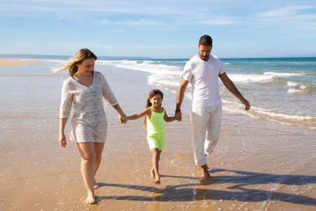Positive relaxed family couple and little girl walking on wet golden sand on beach, kid holding parents hands. Front view. Family outdoor activities or leisure time at sea conceptの写真素材