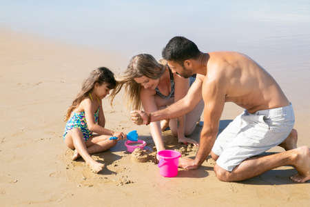 Young parents and sweet little girl playing together on beach, building sand constructions with toy shovel, bucket and bowl. Childhood and family outdoor activity conceptの写真素材