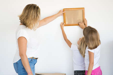 Mother and daughters hanging blank photo frame on white wall. Back view of blonde mom holding and...