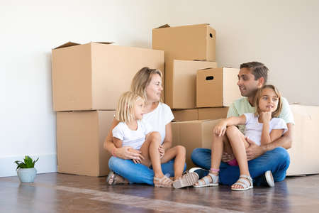 Happy Caucasian family sitting on floor with children. Mother, father and two daughters surrounded...
