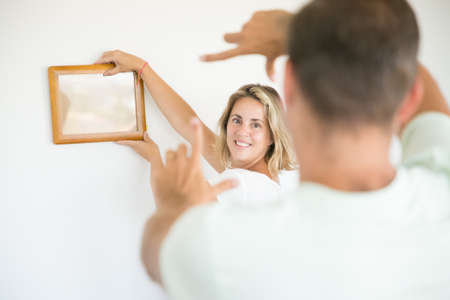 Caucasian couple hanging picture on wall in their new house. Smiling pretty blonde woman holding frame with copy space and man helping her with aligning. Family, relocation and moving day conceptの写真素材