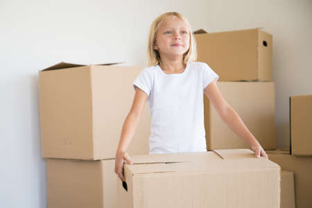 Portrait of adorable girl standing with carton box and looking away. Cute little female kid holding cardboard box during removal in new home. Real estate, relocation and moving day conceptの写真素材