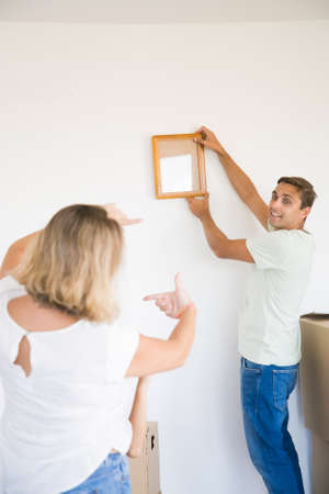 Happy man in casual clothes hanging up frame with copy space on wall in new home. Back view of blonde woman helping him with aligning empty picture. Family, relocation and moving day conceptの写真素材