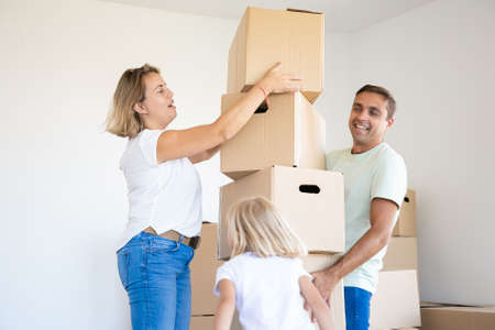 Happy father holding stack of boxes and smiling. Blonde mother in jeans helping him. Daughter...