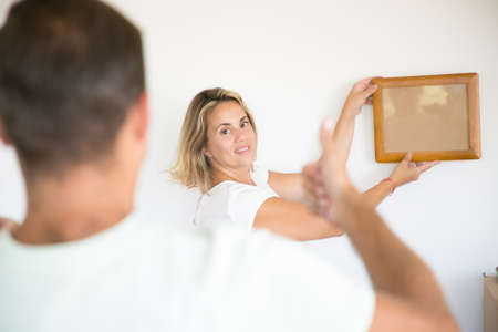 Smiling attractive woman holding and putting frame on wall. Unrecognizable man thumbing up and helping with aligning of picture. Couple decorating new home. Family, relocation and moving day conceptの写真素材
