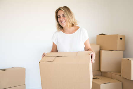 Blonde Caucasian woman carrying carton box in new house or apartment. Happy beautiful lady standing...