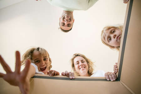 Caucasian family with cute kids looking inside cardboard box. Happy blond mom, dad and two daughters opening box and smiling. Bottom view. Unpacking, relocation and moving day conceptの写真素材