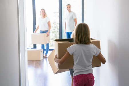 Back view of blonde girl carrying cardboard box and entering into room of new apartment or house. Parents in jeans standing inside with carton boxes and smiling. Relocation and moving day conceptの写真素材