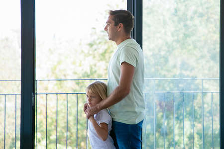 Happy dad embracing daughter and looking in panoramic window. Caucasian father with adorable girl standing near balcony of new apartment or house. Mortgage, relocation and moving day conceptの写真素材