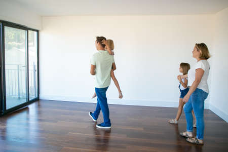 Side view of dad holding daughter and going to balcony with her. Pretty blonde mother in casual clothes standing with another daughter, holding her hand and smiling. Relocation and moving day conceptの写真素材