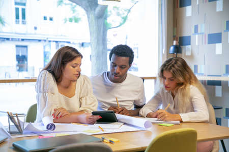 Creative team working on design project together. Professionals drawing in blueprints, writing notes in notebook, watching content on tablet. Medium shot. Creative job and teamwork conceptの写真素材