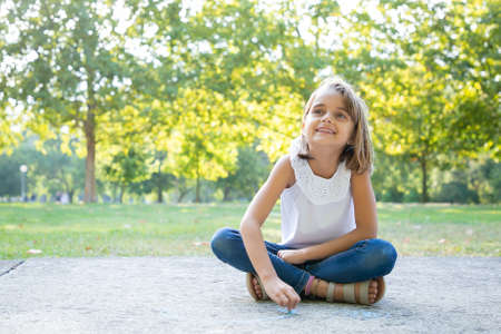 Cheerful pretty girl sitting and drawing with colorful pieces of chalks, looking away and smiling. Front view. Childhood and creativity conceptの写真素材