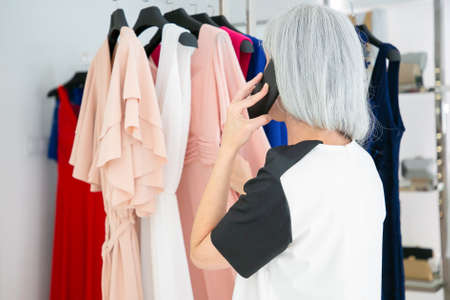Blonde woman talking on mobile phone while choosing clothes and browsing dresses on rack in fashion...