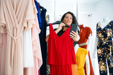 Exited woman shopping in clothes store and consulting friend on cellphone, showing red dress on hanger at frontal camera. Medium shot. Boutique customer or communication conceptの写真素材