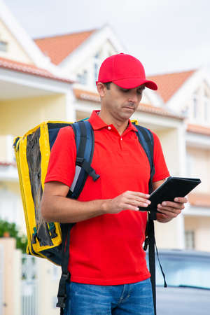 Caucasian courier standing and reading address in tablet. Focused deliveryman delivering express...