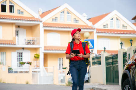 Latin deliverywoman looking for address and holding tablet. Young female courier delivering order in yellow thermo backpack. Houses on background. Delivery service and online shopping conceptの写真素材