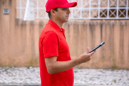 Thoughtful man holding tablet, standing and looking away. Cropped side view of Caucasian courier in red uniform looking for required address. Selective focus. Delivery service and post conceptの写真素材