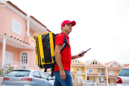 Confident courier going on road, delivering order and working on post. Middle-aged deliveryman wearing jeans, red cap and shirt, carrying yellow thermal backpack. Delivery service and post conceptの写真素材