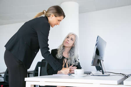 Project manager affixing signature on employees report. Female business colleagues sitting and standing at workplace with monitor and coffee cup. Business communication conceptの写真素材