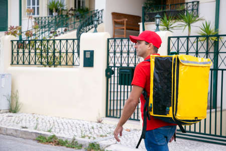 Serious deliveryman looking for address and carrying yellow thermal bag. Attractive courier in red...