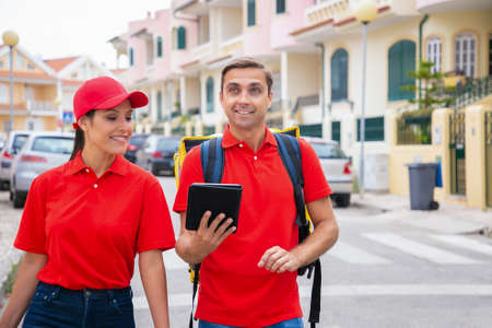 Cheerful post office workers looking for address on tablet. Man carrying yellow thermal bag....