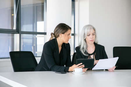 Focused professionals analyzing reports together. Two businesswomen sitting together, reading documents, using tablet and talking. Teamwork conceptの写真素材