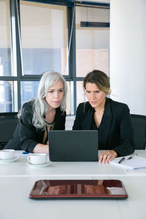 Focused excited businesswomen looking at laptop display while sitting at table with cups of coffee...