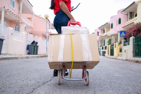 Unrecognizable courier delivering order and walking with boxes on trolley. Cropped deliverywoman holding tablet and looking for required address. Delivery service and online shopping conceptの写真素材