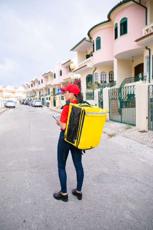 Female post worker in red cap standing with yellow thermal bag and holding tablet. Deliverywoman...