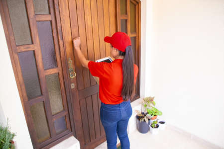 Latin courier knocking on door, holding tablet and carton boxes. Brunette long-haired deliverywoman in red uniform standing in front of door and delivering order. Delivery service and post conceptの写真素材