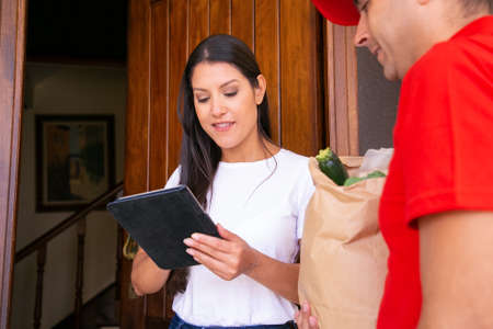 Serious female client watching order details on tablet and receiving order from grocery store....