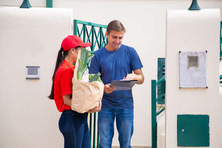 Focused Caucasian man standing and signing in order sheet. Brunette female courier in red uniform...
