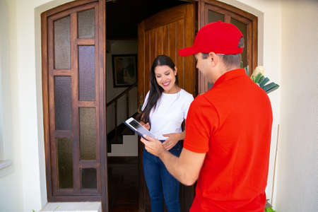Smiling female customer standing near open door and receiving order from grocery store. Caucasian courier delivering order and holding tablet with address. Food delivery service and shopping conceptの写真素材