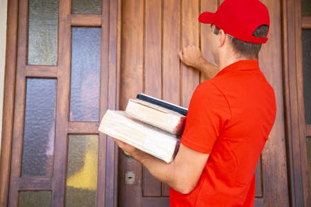 Confident male courier delivering order and knocking on door. Caucasian deliveryman wearing red cap...
