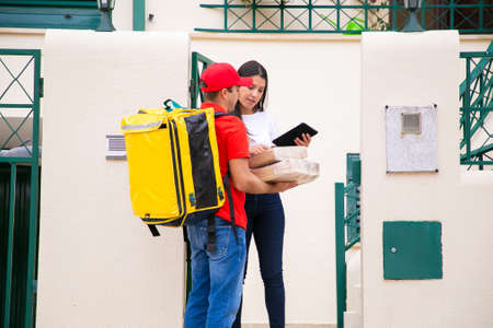 Serious deliveryman holding parcels and woman checking order. Content courier in red cap and shirt with yellow thermal bag delivering express order on foot. Delivery service and post conceptの写真素材