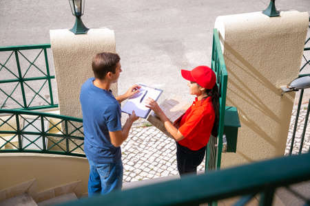 Happy female post worker delivering order to man at home. Caucasian male customer standing,...