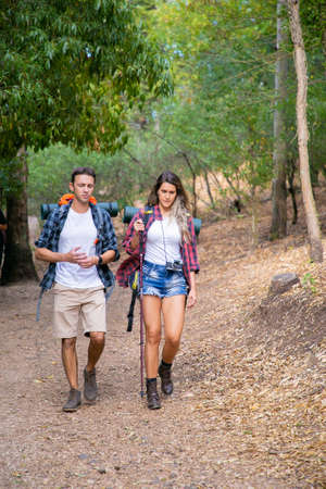 Two young hikers with hiking poles walking on road of mountain forest. Caucasian couple carrying...