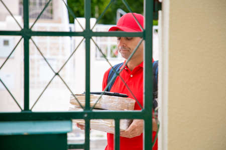 Pensive courier in uniform carrying parcels to customers door, looking at tablet on stack of boxes....