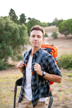 Portrait of pensive male traveler standing on nature and looking at camera. Handsome Caucasian man traveling and carrying backpack. Backpacking tourism, adventure and summer vacation conceptの写真素材