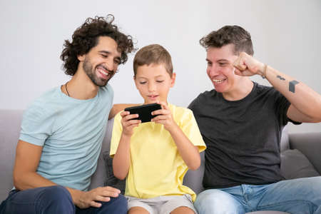 Excited boy playing game on mobile phone, his two happy dads sitting near him and helping. Front...