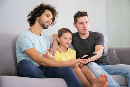 Excited homosexual dads and son watching TV show at home, sitting on couch in living room, hugging,...