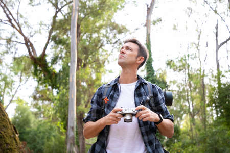 Pensive guy holding camera, looking away and choosing shoot. Caucasian backpacker exploring nature...
