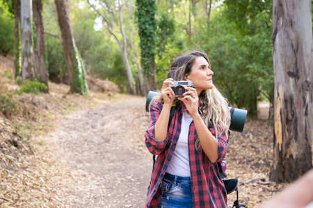 Pensive lady holding camera, looking around and standing on road. Female tourist exploring nature...