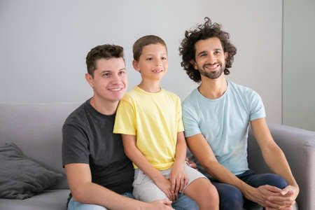 Happy gay fathers and child sitting on couch at home, smiling, and looking away. Front view. Family...
